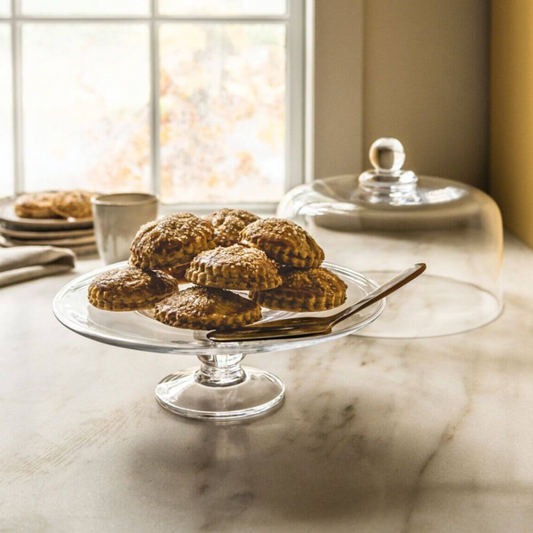 The Simon Pearce Hartland Cakeplate, expertly crafted by glassblowers, displays golden-brown pastries with a knife nearby on a marble counter. Sunlight streams through the window as its glass dome rests to the side.