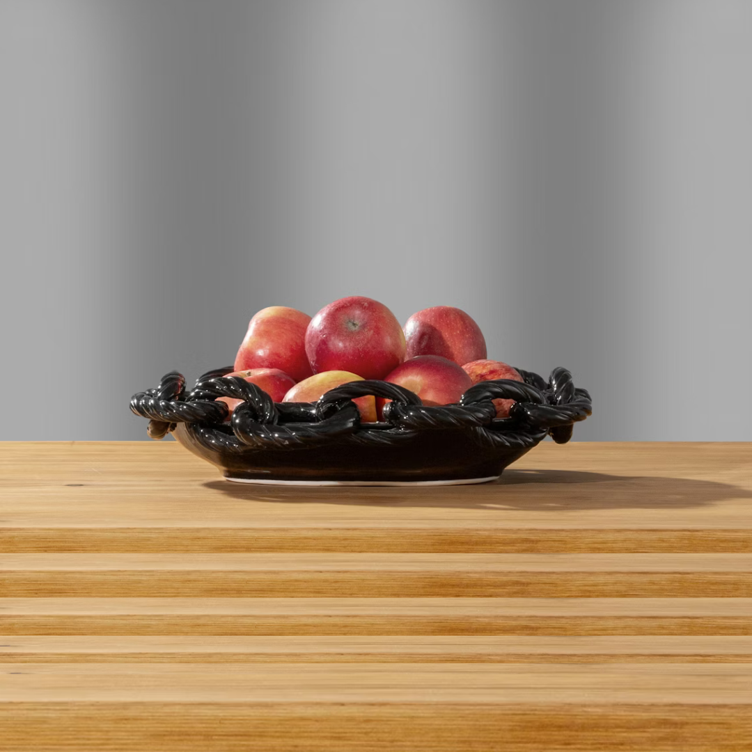 The Schwung Design Link Round Bowl - Black, ideal as serveware or sculptural décor, holds red apples atop a light wooden table with a plain gray background.
