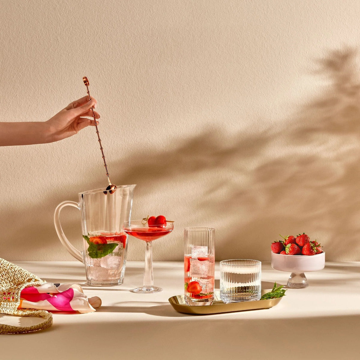 A hand stirs a glass pitcher of pink drink with ice, mint, and berries on a table set with Nude Glassware Big Top Coupe Glasses Set of 2, a bowl of strawberries, and a gold tray in soft sunlight with leafy shadows on the wall.