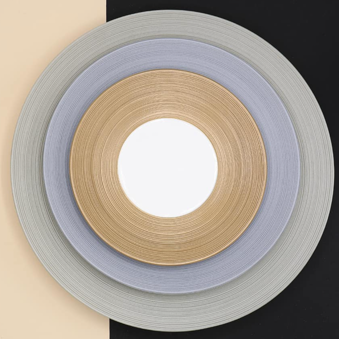 Three concentric circular mats in gold, silver, and gray—evoking JL Coquet’s Hemisphere Dinnerware Grey—are centered over a diagonal beige and black background with a white center.