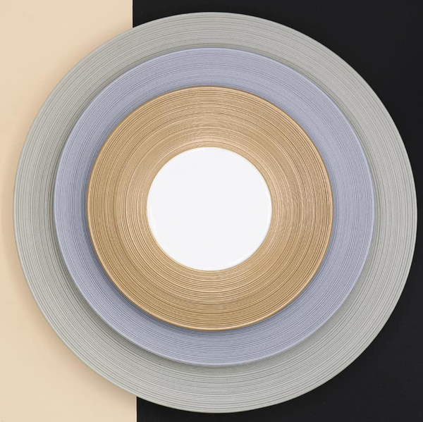 Three concentric circular mats in gold, silver, and gray—evoking JL Coquet’s Hemisphere Dinnerware Grey—are centered over a diagonal beige and black background with a white center.