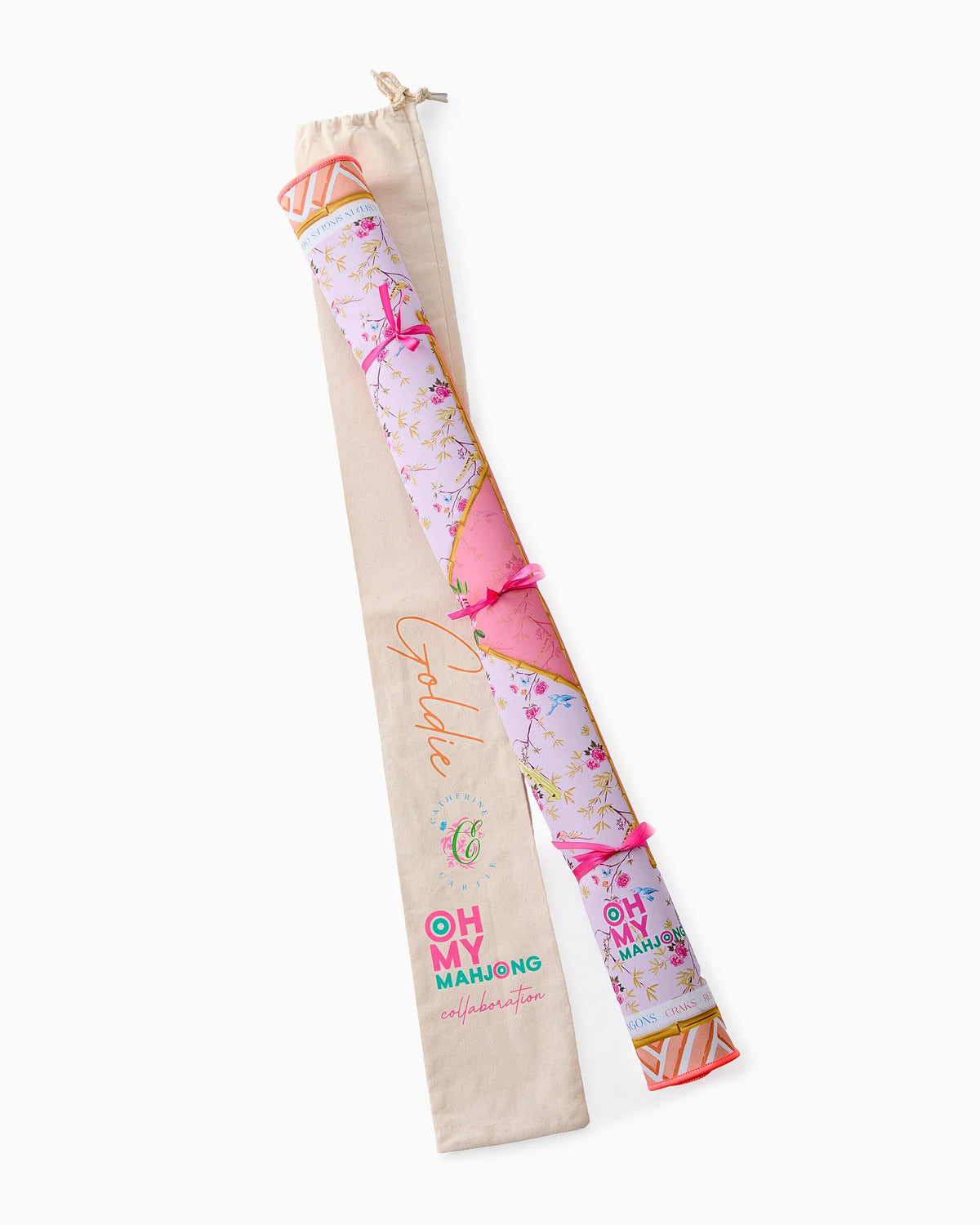 A pink, floral-patterned rolled Mahjong mat from Oh My Mahjong, tied with pink bows, sits beside a cream fabric carrying bag labeled as part of the Golde x Oh My Mahjong collaboration.