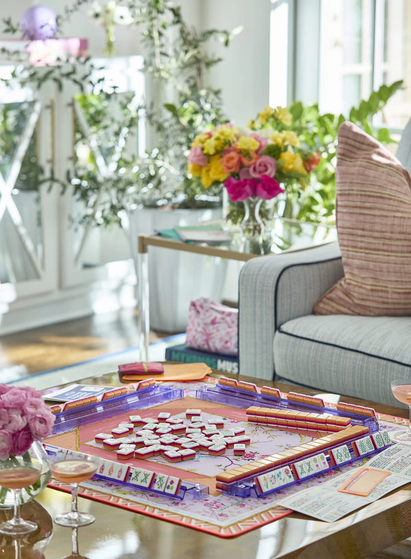An Oh My Mahjong Mahjong Mats set is displayed with floral tiles, two pink cocktails, and a bouquet of flowers in the background.