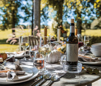 An elegant outdoor table set for a meal features a Simon Pearce Echo Lake Wine Coaster holding a wine bottle, with filled glasses, plates, cutlery, napkins, and decorative pumpkins as Vermont sunlight filters through the trees.