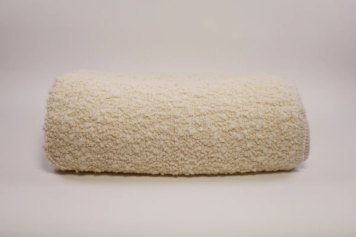 The Uniquity Kusama Boucle Cream Throw, featuring a fluffy cream texture, is styled on a plain light surface with a neutral backdrop, evoking luxurious Belgian craftsmanship.