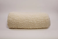 The Uniquity Kusama Boucle Cream Throw, featuring a fluffy cream texture, is styled on a plain light surface with a neutral backdrop, evoking luxurious Belgian craftsmanship.