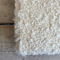 A close-up of the Kusama Boucle Cream Throw by Uniquity, featuring a textured, cream boucle finish, shown draped over a light wooden floor.