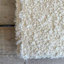 A close-up of the Kusama Boucle Cream Throw by Uniquity, featuring a textured, cream boucle finish, shown draped over a light wooden floor.