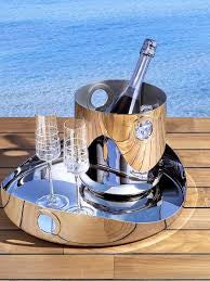 A Christofle Oh De Stainless Steel Round Tray holds a champagne bottle in an ice bucket and two flutes on a wooden table, ideal for outdoor entertaining with the blue ocean in the background.