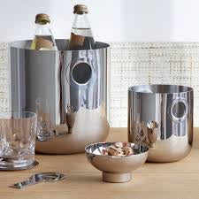 A modern barware set featuring the Christofle Oh De Stainless Steel Champagne Bucket holding two bottles, a matching container, snack bowl, glass with ice, and bottle opener—all displayed on a light wooden surface.