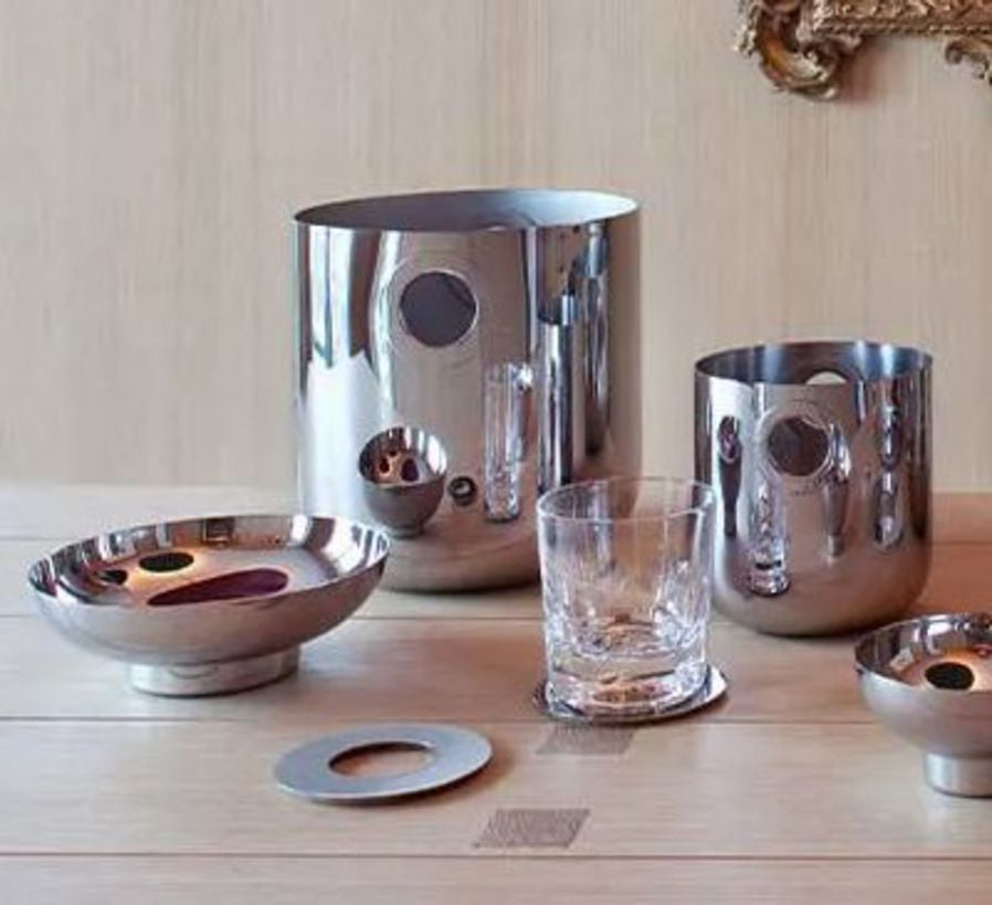 Christofle’s Oh De Stainless Steel Champagne Bucket is arranged on a light wooden table against a beige wall, accompanied by barware essentials and décor featuring a gold frame in the corner.