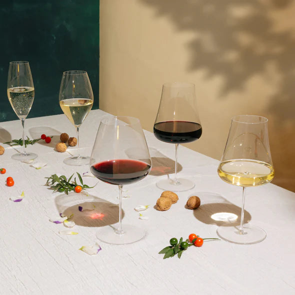 Five Nude Glassware Stem Zero Elegant Red Wine Glasses perch elegantly on a white table, their thin stems and crystal cut edges catching the light. Nuts, petals, and small berries are scattered artfully around them, while shadows play across colored backgrounds in the scene.