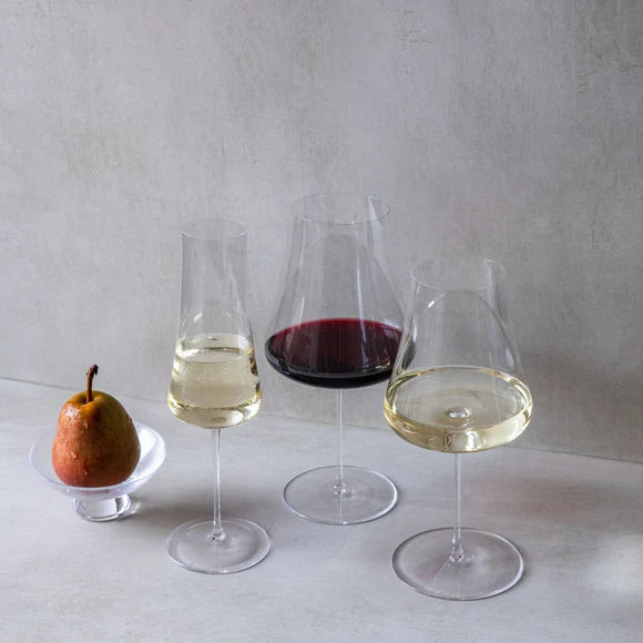 A pear rests on a glass dish beside three wine glasses—two with slender stems filled with white wine and one Stem Zero Elegant Red Wine Glass by Nude Glassware—gracefully arranged on a light surface against a plain background. Each glass features a subtle crystal cut edge, lending an extra element of sophistication to the scene.