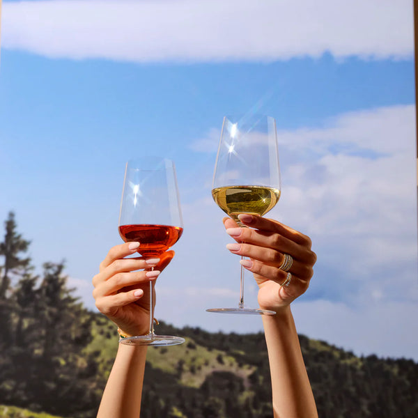 Two manicured hands hold up a Nude Glassware Stem Zero Delicate White Wine Glass and a filled wine glass, set against blue sky, clouds, and green hills.