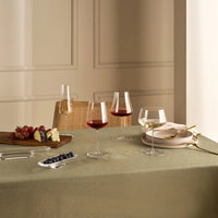 A dining table set with a light green tablecloth features the Nude Glassware Stem Zero Powerful Red Wine Glass filled with red wine, a champagne flute, plates with gold cutlery, and a wooden cheese board topped with grapes and berries. A chair is in the background.