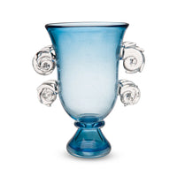 The Yakush Sal Blue Vase is a hand-blown glass vase featuring a wide mouth, narrow base, and four clear, spiral seashell-inspired handles evenly spaced around its blue body.