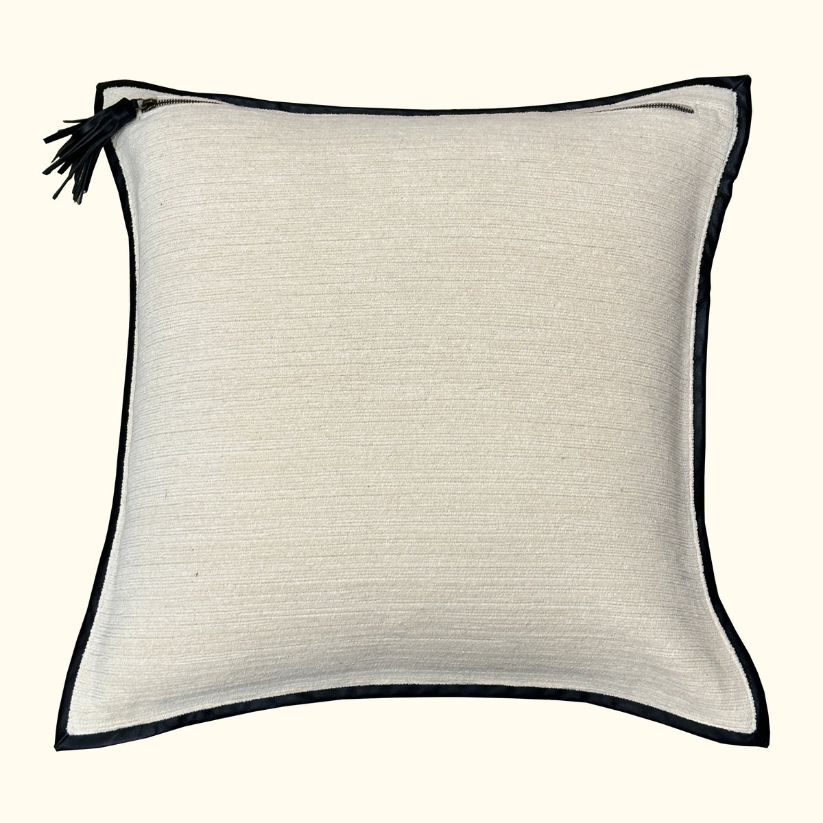 The Leather Frame Pillow Ivory by Threads Soft Goods is a square, ivory accent pillow with black piping and a small black tassel on one corner, shown against a plain background.
