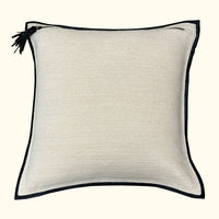 The Leather Frame Pillow Ivory by Threads Soft Goods is a square, ivory accent pillow with black piping and a small black tassel on one corner, shown against a plain background.