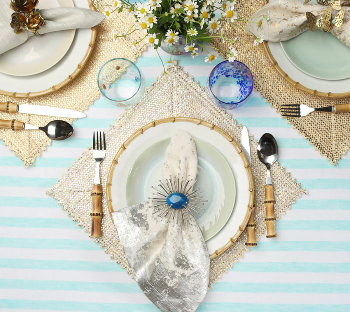 Three place settings on a striped table, each featuring a textured square Kim Seybert Stamped Reversible Placemat Silver & Gold, dinner plates, bamboo-handled silverware, and a napkin held by a decorative ring.