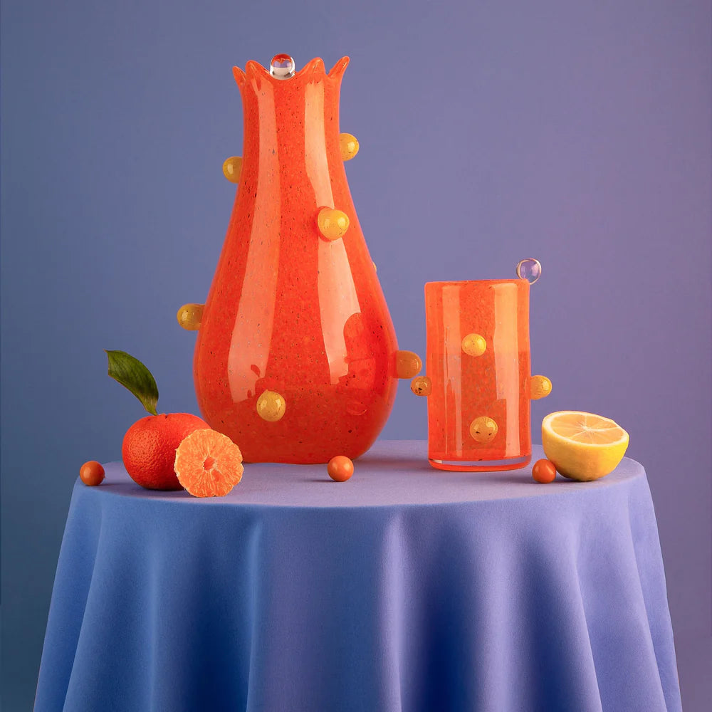 The Yakush Pollen Bloom Vase - Orange and matching glass with yellow knobs rest on a blue cloth with a tangerine, lemon half, and orange balls, set against a purple background.