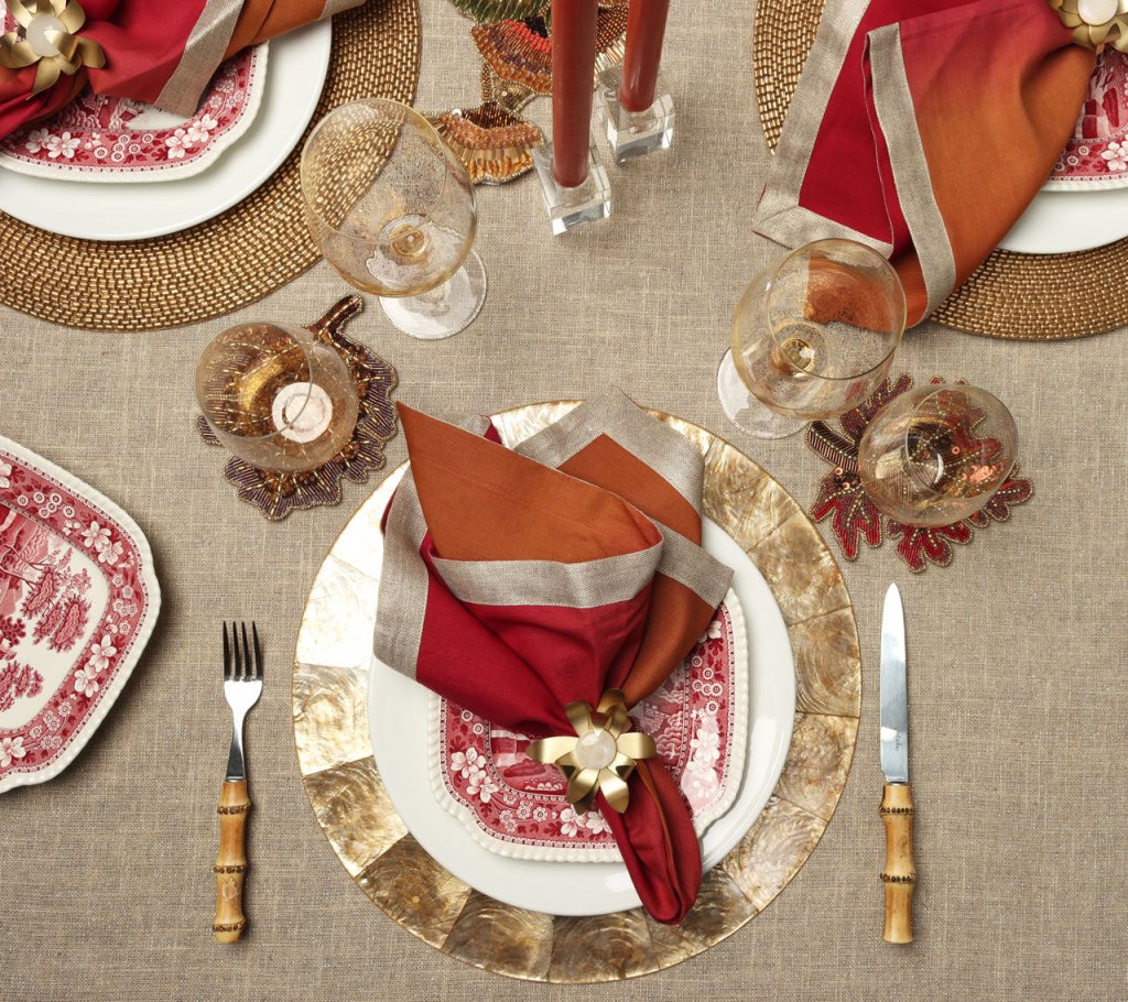 Elegant Thanksgiving table set with Kim Seybert’s Fringe Tablecloth in Natural & Silver, gold-rimmed plates, red/orange napkins with gold rings, vintage red dishes, wine glasses, autumn leaf decor, and bamboo-handled cutlery.