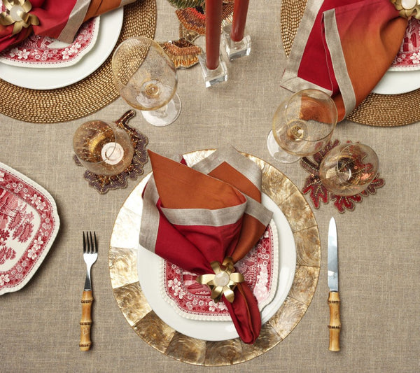 Elegant Thanksgiving table set with Kim Seybert’s Fringe Tablecloth in Natural & Silver, gold-rimmed plates, red/orange napkins with gold rings, vintage red dishes, wine glasses, autumn leaf decor, and bamboo-handled cutlery.