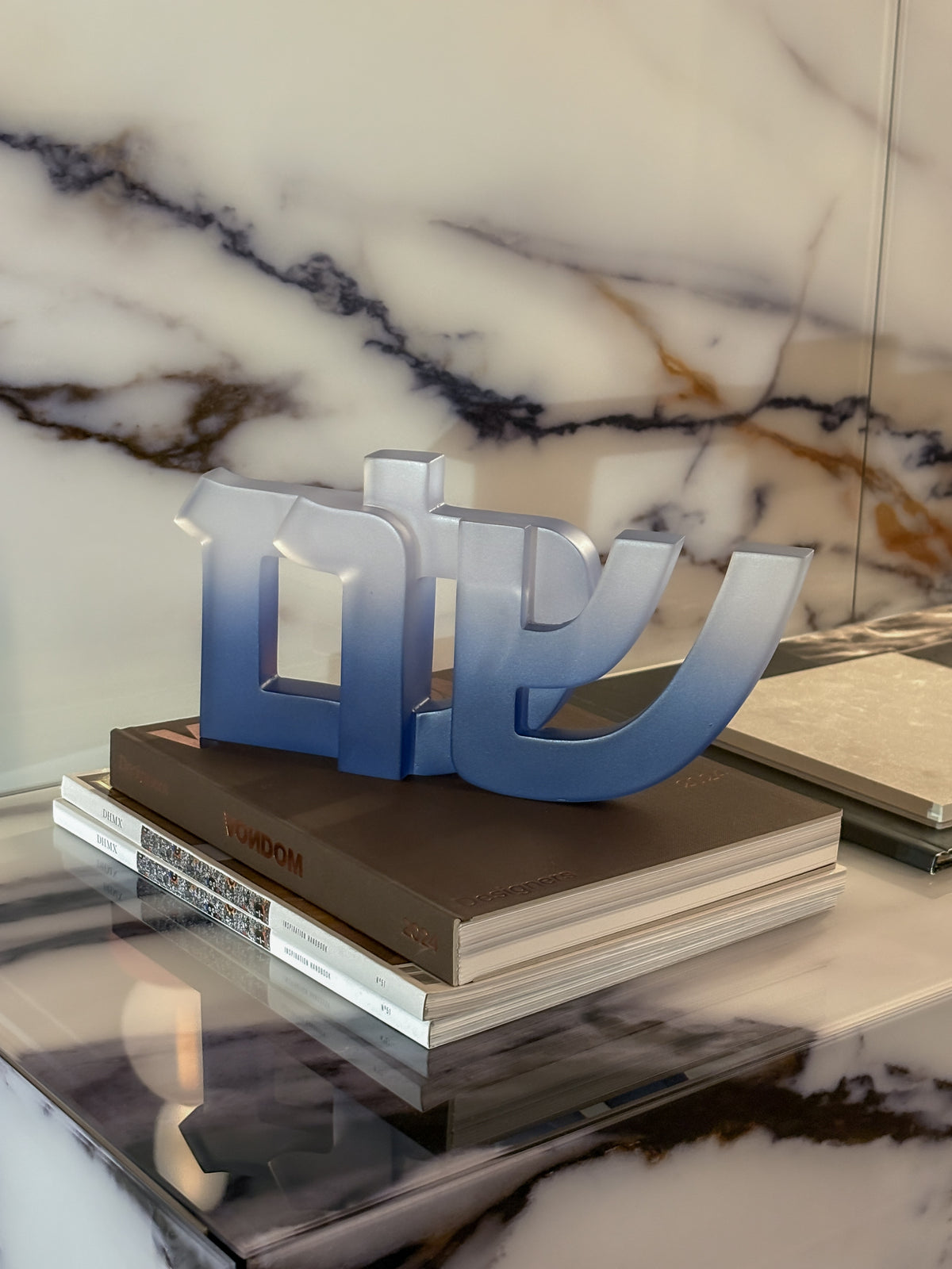 The Shalom Sculpture by CDMX / BRUCI, featuring Hebrew letters in gradient blue resin, sits atop stacked coffee table books on a marble surface with a marbled backdrop—bringing modern Jewish home decor charm.