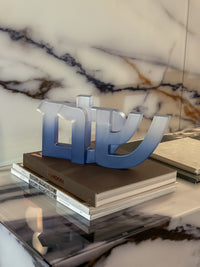 The Shalom Sculpture by CDMX / BRUCI, featuring Hebrew letters in gradient blue resin, sits atop stacked coffee table books on a marble surface with a marbled backdrop—bringing modern Jewish home decor charm.