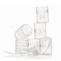 Six clear borosilicate glass tumblers from the Deco Clear Water Glass Set of 6 by Ichendorf Milano, each featuring distinctive etched or molded designs, are stacked atop one another. A white ribbon winds through and around the glasses, elegantly draping across their surfaces. The background is a clean white.
