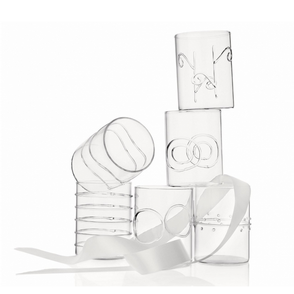 Six clear borosilicate glass tumblers from the Deco Clear Water Glass Set of 6 by Ichendorf Milano, each featuring distinctive etched or molded designs, are stacked atop one another. A white ribbon winds through and around the glasses, elegantly draping across their surfaces. The background is a clean white.
