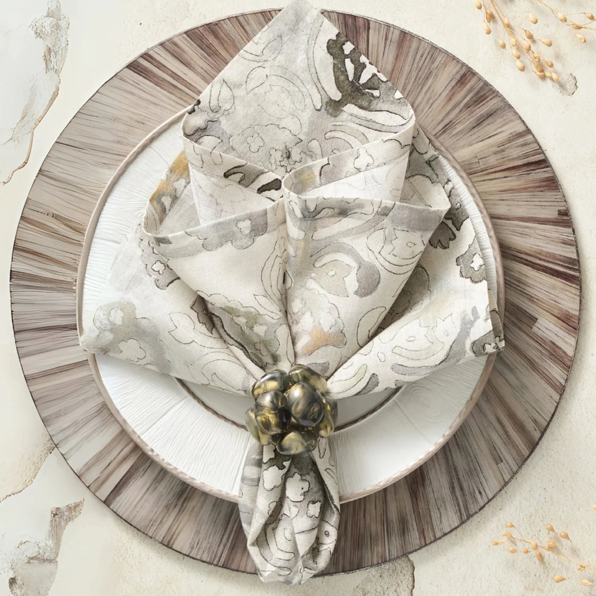 A decorative table setting features a patterned beige napkin from the Kim Seybert Giverny Napkin Set of 4 in Natural & Grey, elegantly folded with an ornate ring on white and brown-rimmed plates, accented by nearby dried grasses.