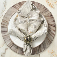 A decorative table setting features a patterned beige napkin from the Kim Seybert Giverny Napkin Set of 4 in Natural & Grey, elegantly folded with an ornate ring on white and brown-rimmed plates, accented by nearby dried grasses.