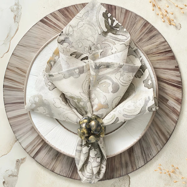 A decorative table setting features a patterned beige napkin from the Kim Seybert Giverny Napkin Set of 4 in Natural & Grey, elegantly folded with an ornate ring on white and brown-rimmed plates, accented by nearby dried grasses.