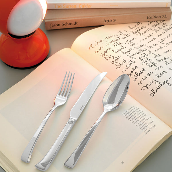 A fork, knife, and spoon from the Sambonet Imagine Flatware 5PPS - Stainless Steel set rest on an open book with handwritten text, near an orange lamp and stacked books in the background.