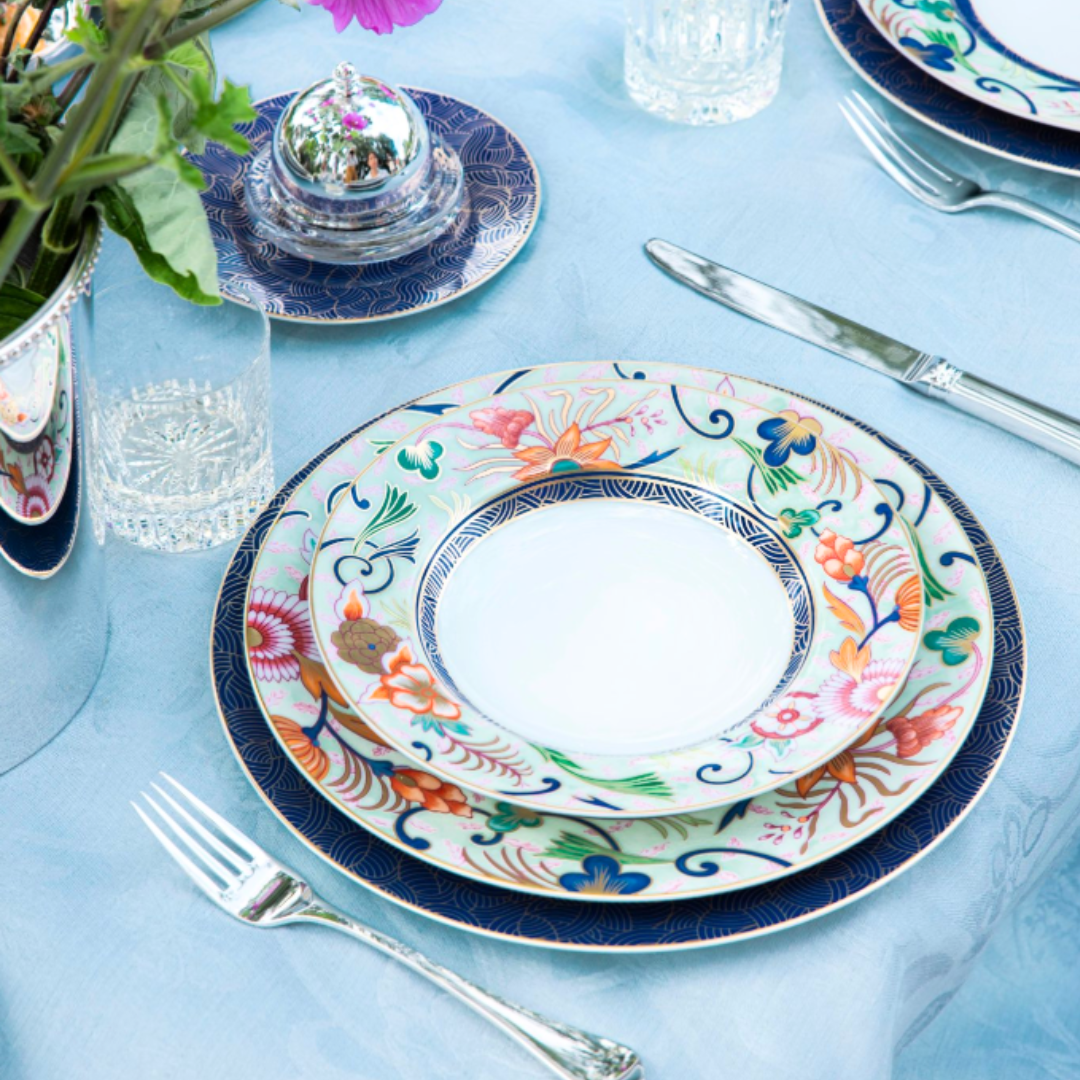 A table setting featuring Raynaud Imari Dinnerware with floral motifs, complemented by silver cutlery, a glass, and a Japanese-inspired flower arrangement on a light blue tablecloth.