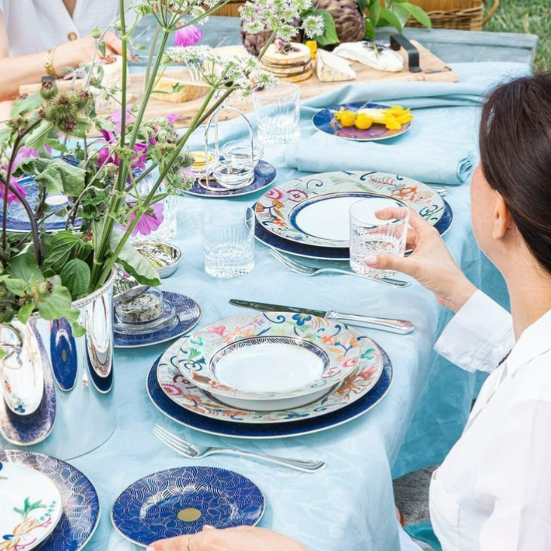 A woman enjoys a meal outdoors with others at a bright summer table set with Raynaud’s Imari Dinnerware, featuring colorful plates inspired by Japanese floral art, alongside glasses and fresh flowers.