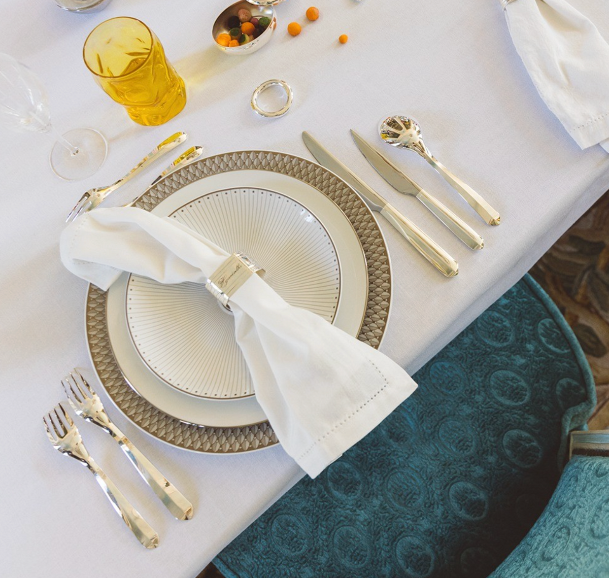 Elegant table set with a white napkin in a silver ring on layered plates, Christofle Infini Flatware 5 Piece Setting in silver plate, an amber glass, and a snack dish on a white tablecloth beside a blue textured chair.