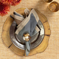 A table setting features a wood-textured charger, silver plate, and a beige and gray napkin held by the Kim Seybert Sea Stone Napkin Ring Beige Set of 4, with two amber glasses and orange flowers on a woven placemat nearby.