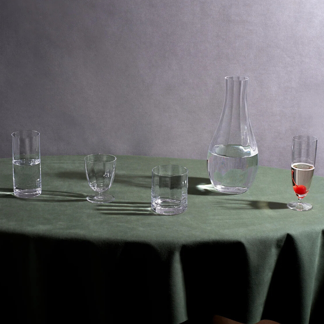 The Iris Drinkware Collection by L'Objet features four mouth-blown glasses and a decanter on a green tablecloth: one glass has water, one is empty, another holds water with a cherry, and the decanter is partly filled with water.