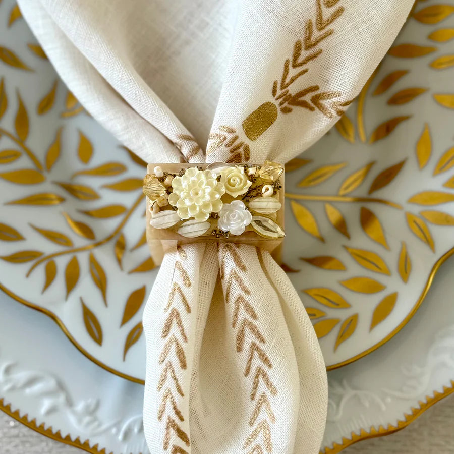 An elegant table decor features a white napkin with a gold leaf pattern, secured by Bodrum’s Ivory Petal Napkin Ring (set of 4), and placed on a white plate adorned with gold leaf designs.