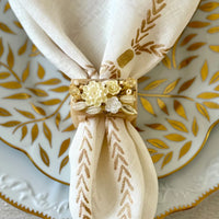 An elegant table decor features a white napkin with a gold leaf pattern, secured by Bodrum’s Ivory Petal Napkin Ring (set of 4), and placed on a white plate adorned with gold leaf designs.