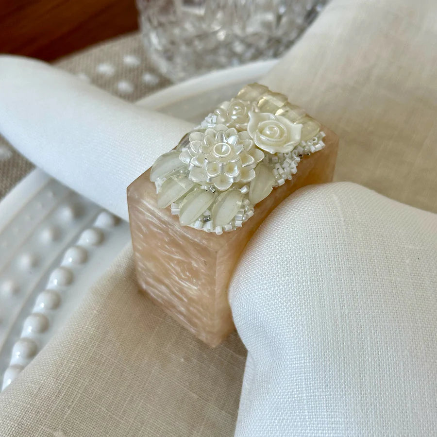 A close-up of the Bodrum Ivory Petal Napkin Ring, featuring intricate ivory floral detailing, holds a white cloth napkin on a set dining table, creating elegant table decor.