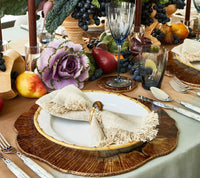 A decorative table setting features a white plate on a wood charger, silver cutlery, glasses, a centerpiece of faux fruits and greens, and a Kim Seybert Fringe Napkin Silver & Natural from the set of 4.
