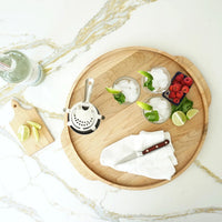 The JK Adams Elmore Round Serving Board displays two iced drinks with lime and mint, a cocktail strainer, a bowl of raspberries and blackberries, and lime wedges. Nearby, a small cutting board holds more lime wedges and a knife on white marble.