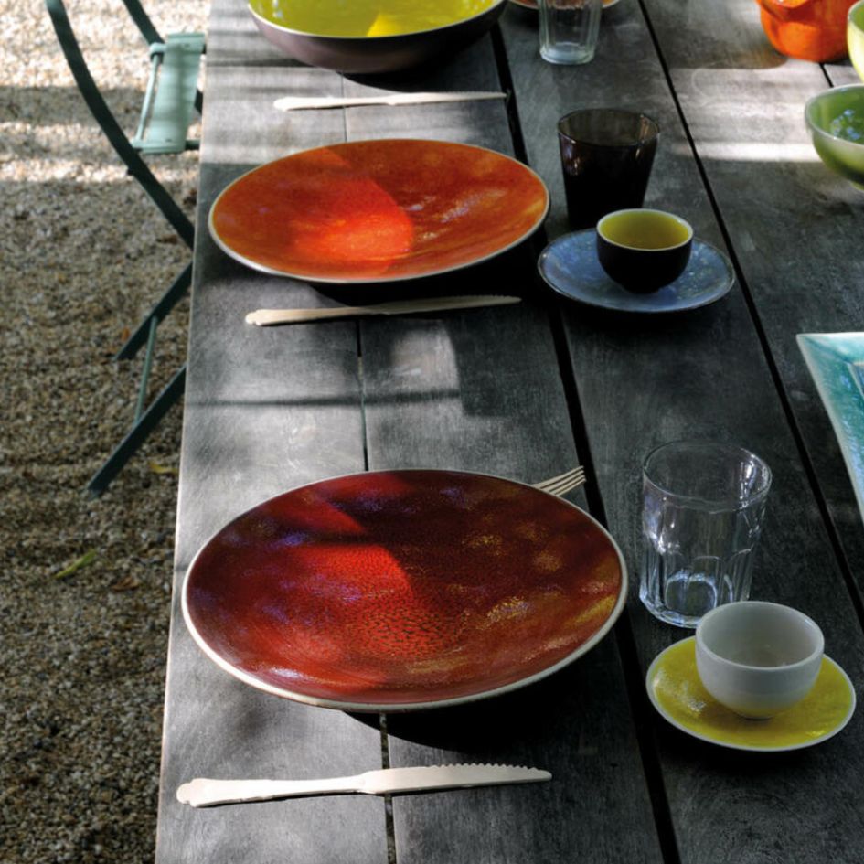 A rustic wooden table is set for a meal with Jars - Rosenthal Tourron Dinnerware Cerise, featuring vibrant ceramic plates in pretty glazes and elegant glassware. Sunlight dapples the inviting scene.