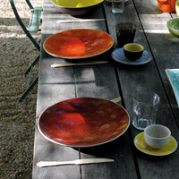A rustic wooden table is set for a meal with Jars - Rosenthal Tourron Dinnerware Cerise, featuring vibrant ceramic plates in pretty glazes and elegant glassware. Sunlight dapples the inviting scene.