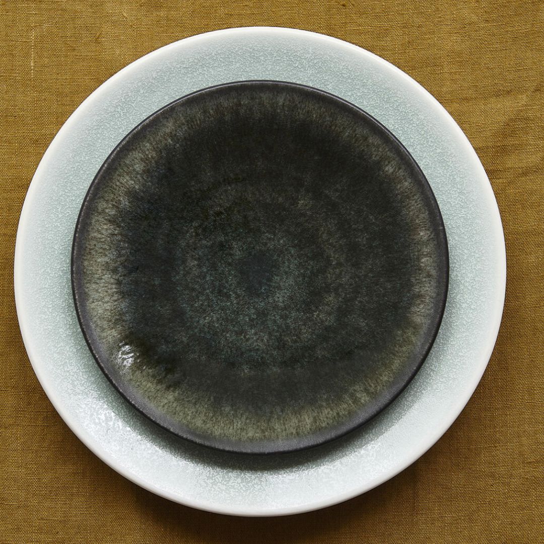 Two ceramic plates from the Jars - Rosenthal Tourron Dinnerware Neige collection are stacked on a brown tablecloth: a light gray, textured-rim plate beneath a smaller plate with a black-to-deep green gradient.