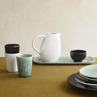 Handmade in France, the Jars - Rosenthal Tourron Dinnerware Neige set features cups, bowls, a white pitcher, and stacked plates on an olive-green tablecloth with a rectangular tray, all set against a neutral background.