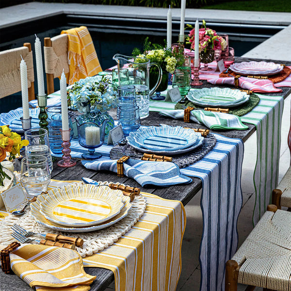 A colorful poolside table showcases Juliska's Cabana Stripe Melamine Dinnerware Set of 4 - Seagrass, with striped runners, matching napkins, elegant glassware, candles, flowers, and bamboo cutlery. The shatterproof plates are dishwasher safe.