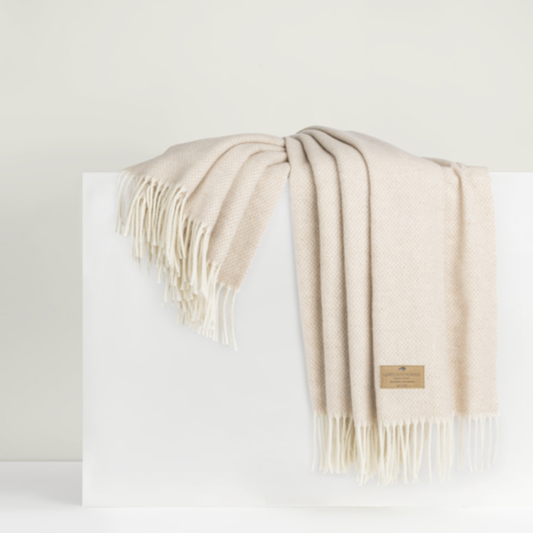 The Lands Downunder Juno Cashmere Throw is a soft, cream-colored luxury blanket with fringe edges and a brown label on one corner, elegantly draped over a white rectangular block.
